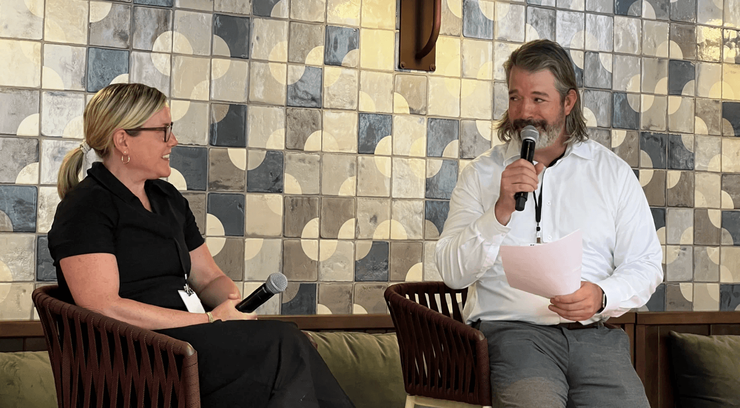 Two people talking on a stage set against a tiled wall, one holding a microphone and notes